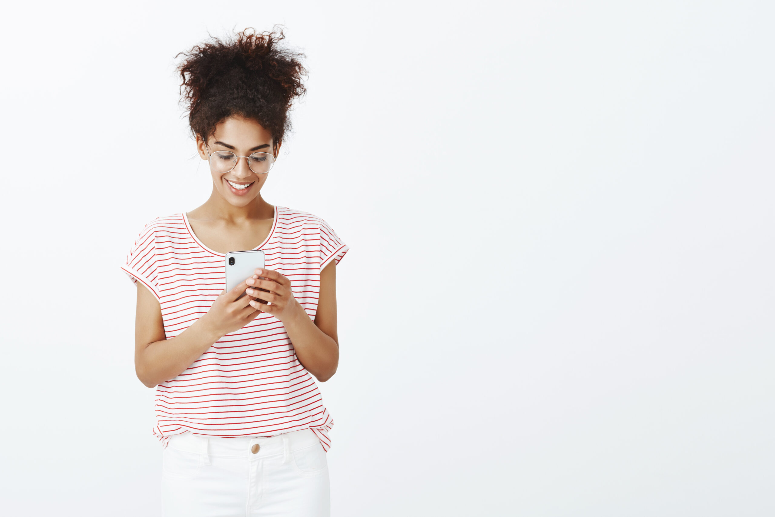 Studio shot of charming carefree young woman with tanned skin in glasses and striped t-shirt, looking and smiling at smartphone screen, receiving pleasant message and replying via device and wifi.